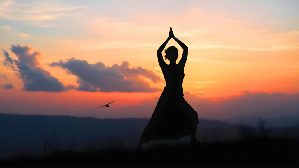 A graceful woman in yoga position on a grassy hill at sunrise, with vibrant clouds and birds in the background.